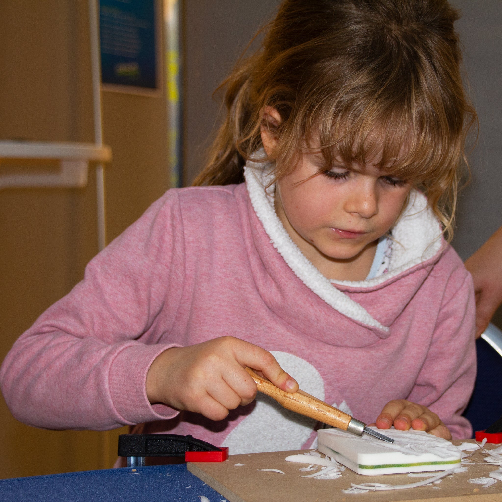 Ein etwa 6-jähriges Mädchen schnitzt mit einem Hohlbeitel einen Block aus Kartoffelholz. Sie ist Anfängerin und lernt das Schnitzen, indem sie das weiße Material vorsichtig vom Block abschnitzt.